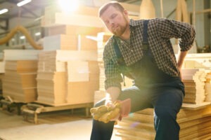 Tired worker with closed eyes sitting on stack of wooden plank and massaging lower back while feeling pain in back